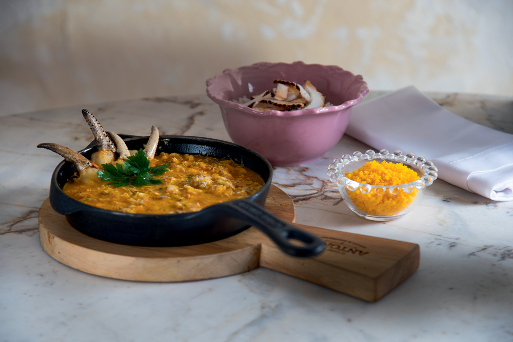 Moqueca servida em panela de ferro sobre suporte de madeira com patinha de caranguejo por cima ao lado de potinho transparente com farofa de dendê e tigela rosada com arroz coberto por fitas de coco.