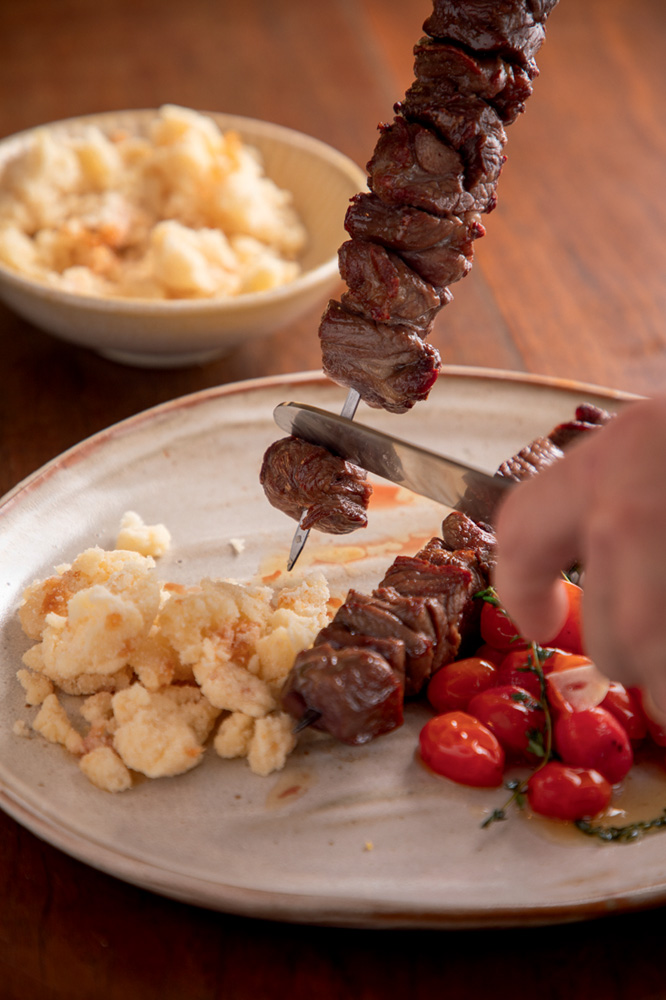 Dia dos Pais – Mocotó Prato de cerâmica farofa, tomates assados e espetinho de carne de sol sendo servido.