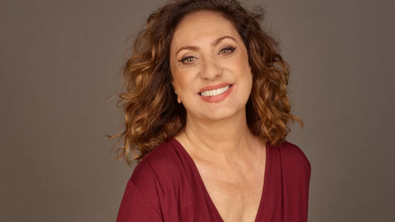 Mulher branca de cabelos encaracolados sorri e veste blusa cor de vinho decotada.