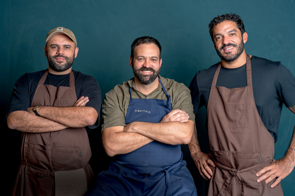 Três homens brancos de barba preta vestidos com avental de cozinha em frente a parede azul.