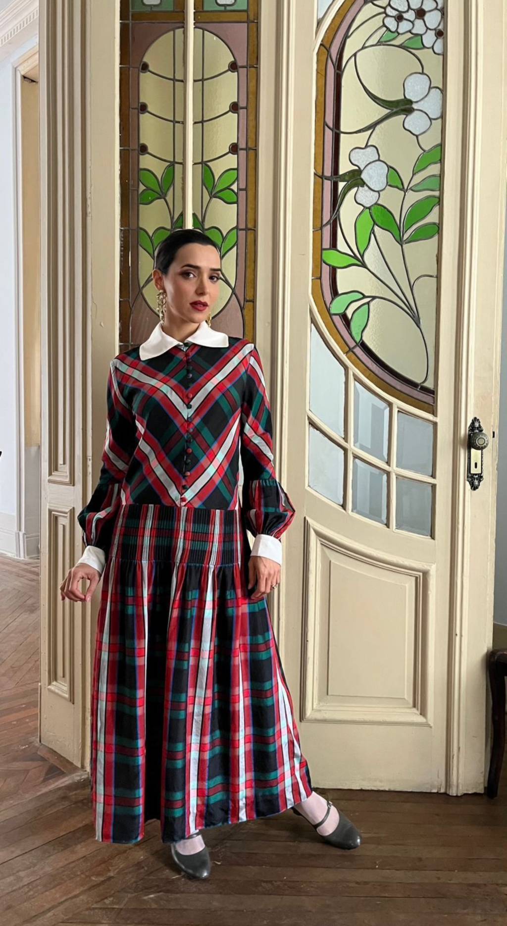 credito Fernanda Cuenca Mulher branca posa com roupa de época, vestido longo, em frente a grande porta de madeira pintada de bege.