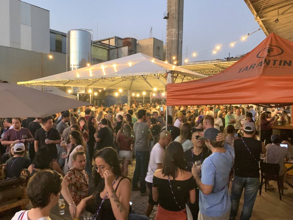 IPA Day SP A imagem mostra um ambiente amplo cheio de pessoas em pé conversando. O local é aberto e tem uma tenda branca e uma vermelha com luzes amarelas em cima.
