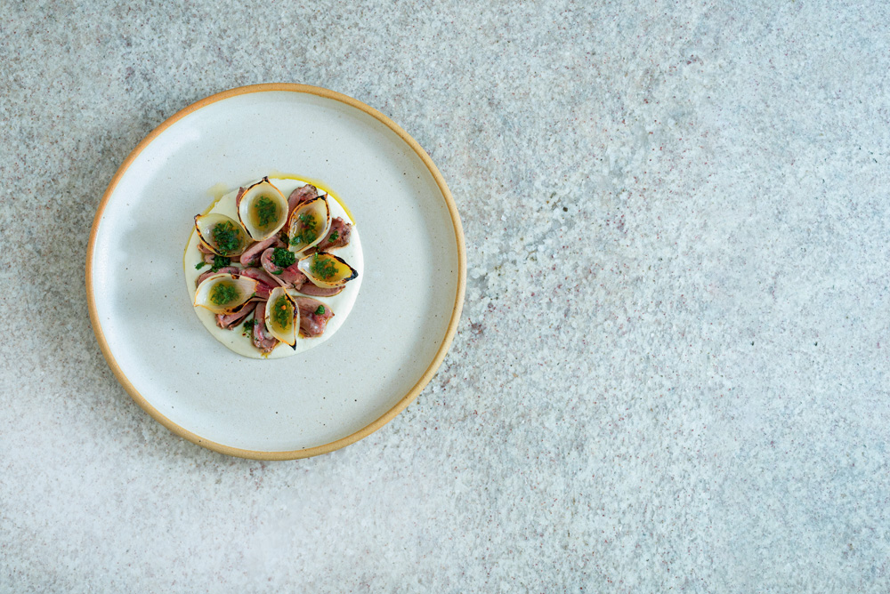 COMER & BEBER 2023 – Cora Foto tirada de cima mostra um prato branco com corações de pato, creme de couve flor e cebola.