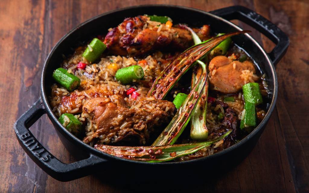 Dalva e Dito A imagem mostra uma panela preta em cima de uma mesa de madeira vista de cima. Dentro, uma mistura de arroz, com quiabos e pedaços de frango.