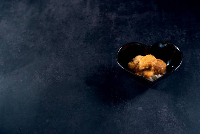 COMER & BEBER 2023 – Murakami Em um fundo preto, um pote preto em formato de coração tem ouriço do mar dentro.