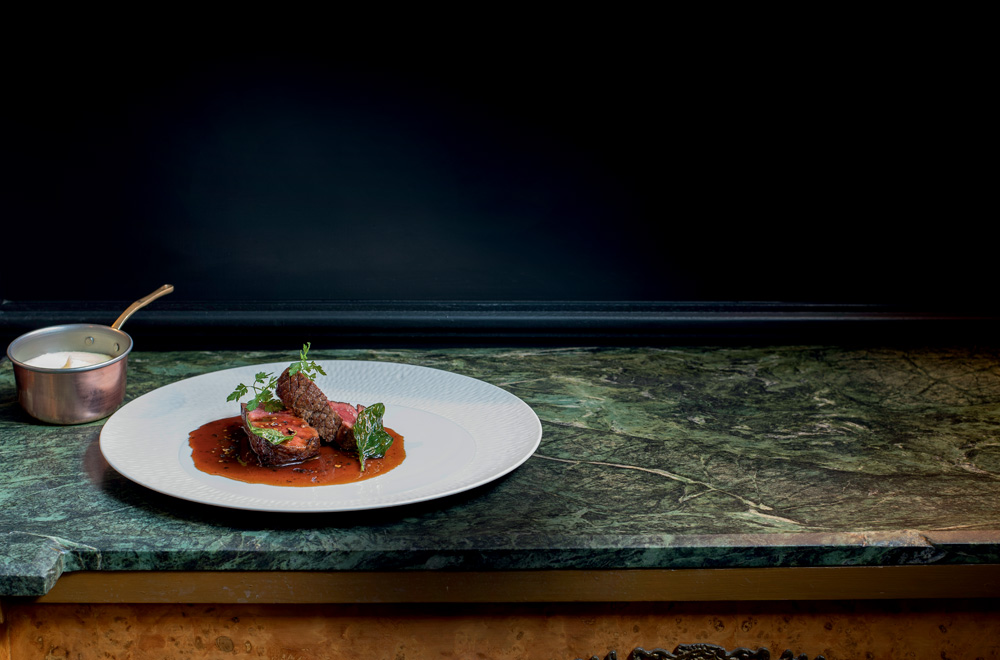 COMER & BEBER 2023 – Président Prato branco com pedaços de carne e molho por cima sobre uma mesa de mármore.
