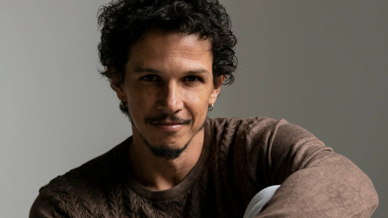 Homem de bigode e cabelos encaracolados dá meio sorriso com mãos apoiadas no joelho. Veste suéter marrom claro.