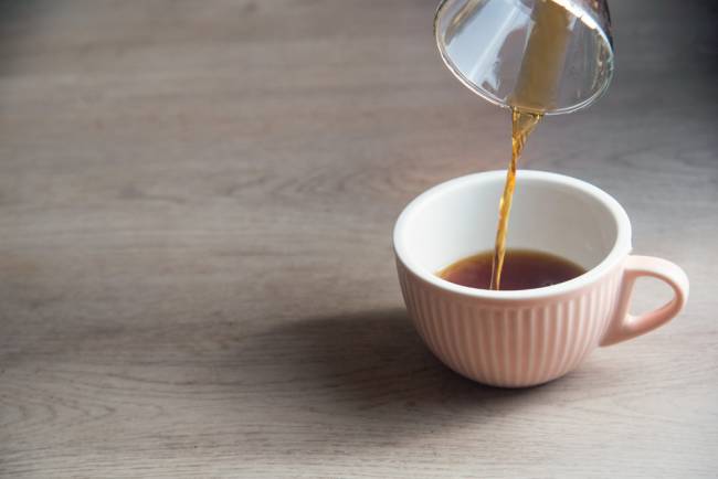 Café coado sendo despejado em uma xícara branca sobre uma mesa de madeira.