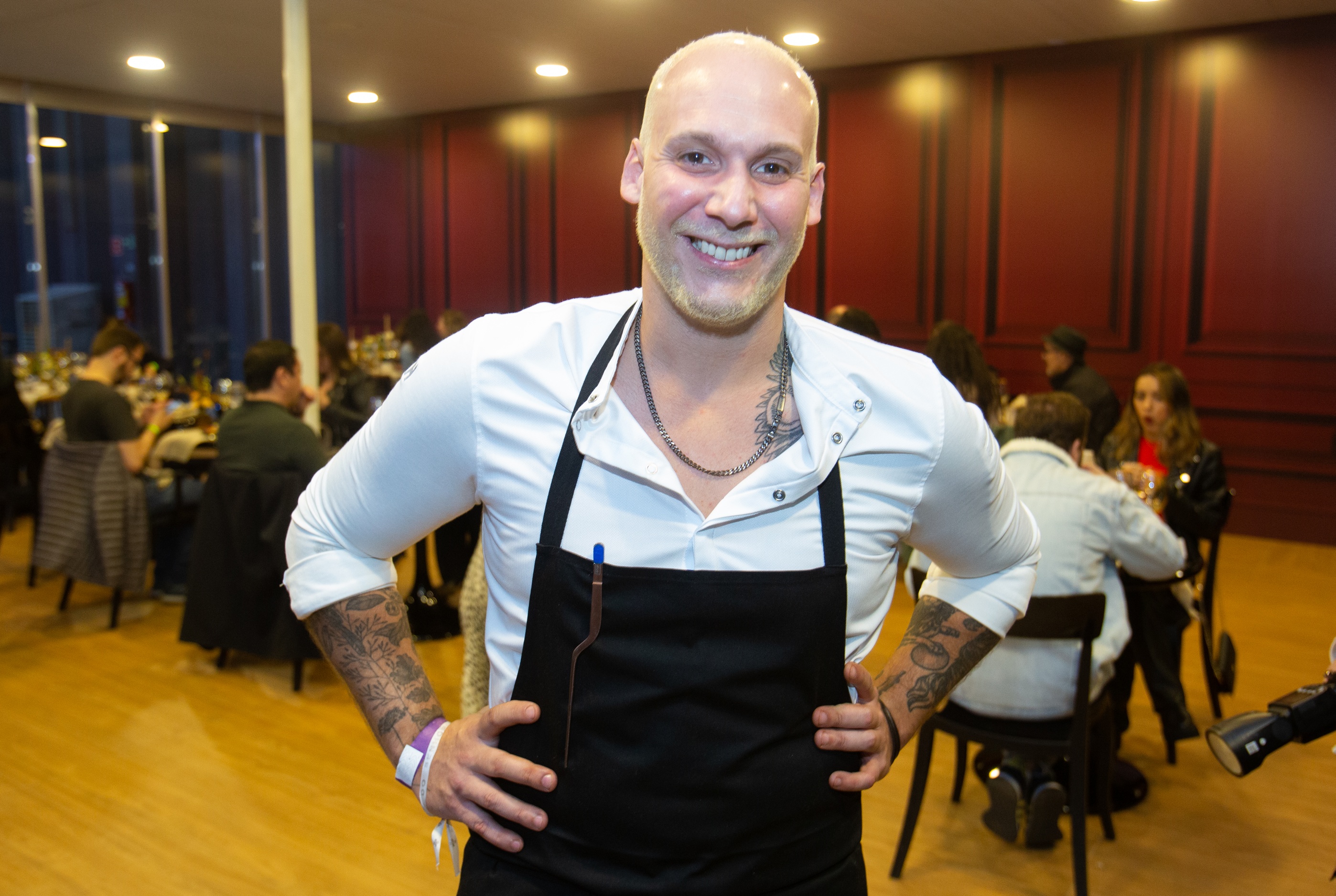 Homem branco sorri para a foto com as m&atilde;os na cintura. Ele usa uma camisa branca e um avental preto.
