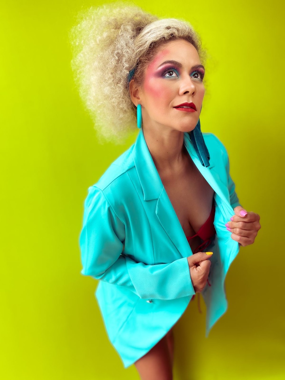 Ariel Moura (Pedro Stkls-Divulgaçação) Mulher de cabelos loiros, quase brancos, posa segurando as lapelas do blazer azul cintilante. Usa brincos e tem maquiagem colorida no rosto. O fundo é todo verde neon.