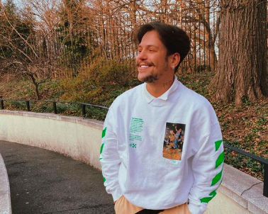 Imagem mostra rapaz sorrindo em parque. Ele usa camisa branca e está com as mão nos bolso de calça e olha para a esquerda da imagem