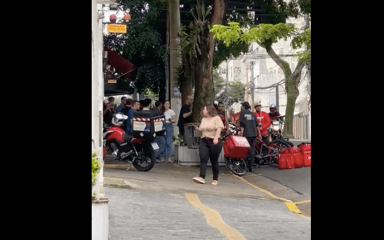 Frame de vídeo mostra pessoas passando na calçada de rua. Ao fundo, parcialmente encoberto por árvores, entregadores discutem com homem branco