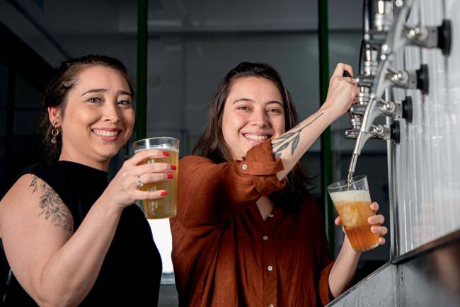 Duas mulheres brancas de cabelos castanhos estão lado a lado sorrindo para a foto. A da esquerda está de camiseta preta e segura um copo com cerveja na mão. A outra segura um copo enquanto abre a torneira que sai cerveja.