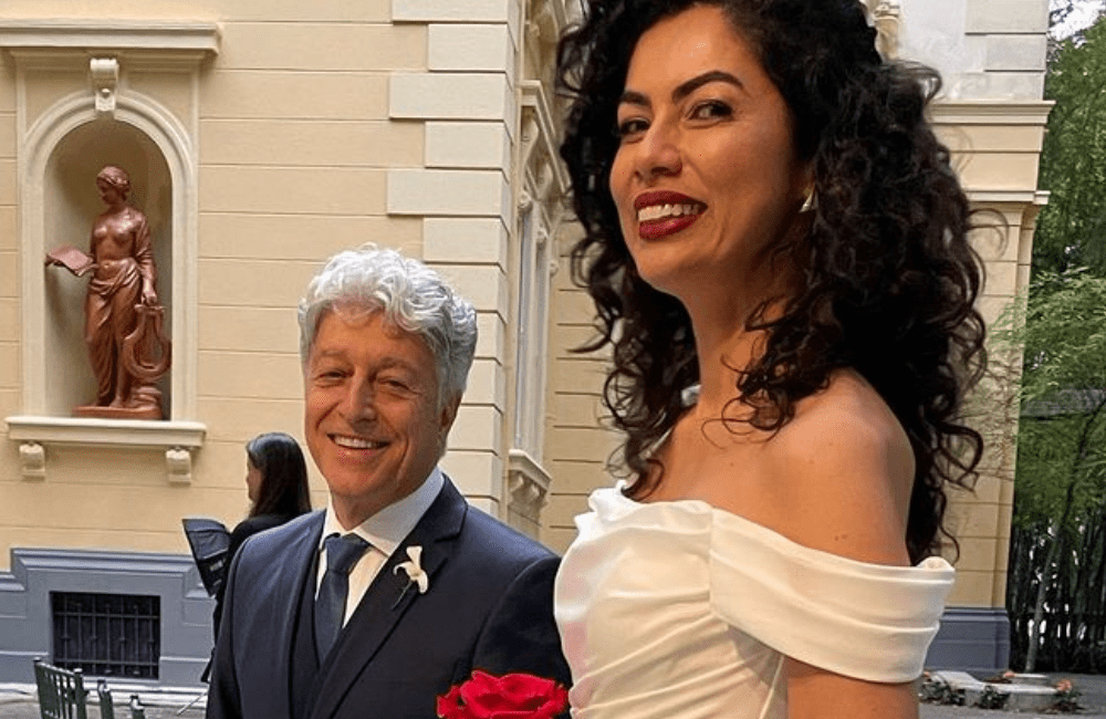 Caco Barcellos usando um terno azul e sorrindo, enquanto a esposa Carla Tilley está ao seu lado, usando um vestido de noiva branco e sorrindo para a camêra