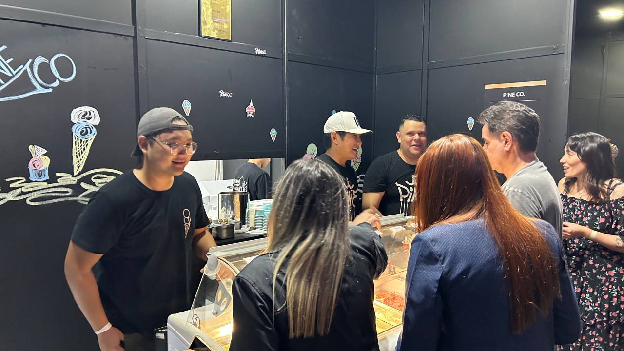 Estande de sorvete em feira com paredes pretas. Pessoas do lado de fora olhando a vitrine de vidro e atendentes de camisa preta do outro lado.