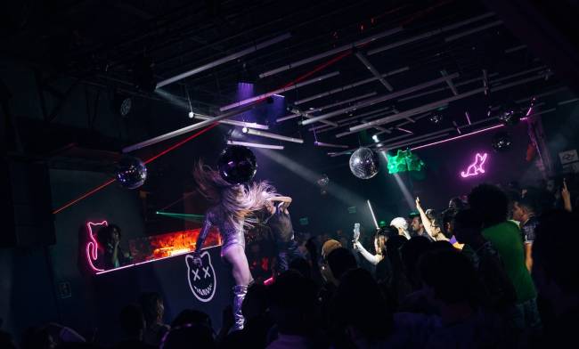 Foto exibe pista escura com plateia de pé e drag queen de peruca prateada dançando no palco.