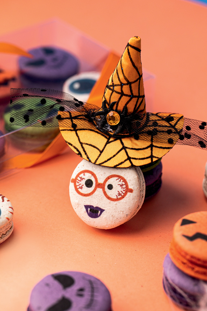 MAYMACARONS-120.jpg Em fundo laranja, um doce com desenho de monstro e chapéu de bruxa.