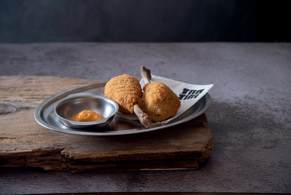 T&aacute;bua de madeira com bandeja de inox coberta com duas coxinhas de frango ao lado de molho alaranjado