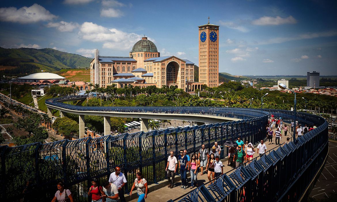 Imagem mostra Santuário de aparecida ao fundo e, em primeiro plano, passarela que leva ao edifício religioso. Paisagem é de dia de céu azul e montanhas ao fundo