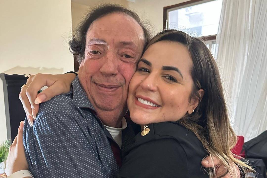 um homem branco, de cabelos pretos, abraçando a ponto de colar o rosto com uma mulher branca de mechas loiras no cabelo. os dois sorriem.