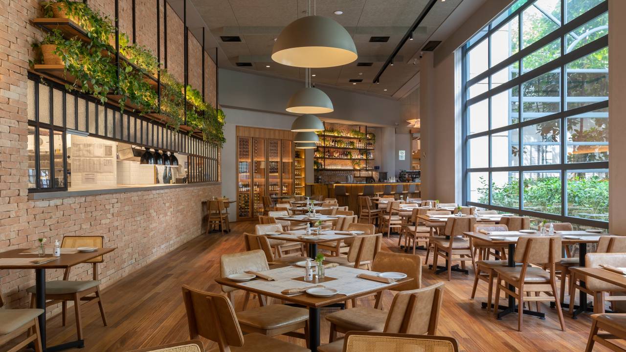 Salão do restaurante Più com mesas de madeira, cozinha aberta à esquerda e janelão à direita.