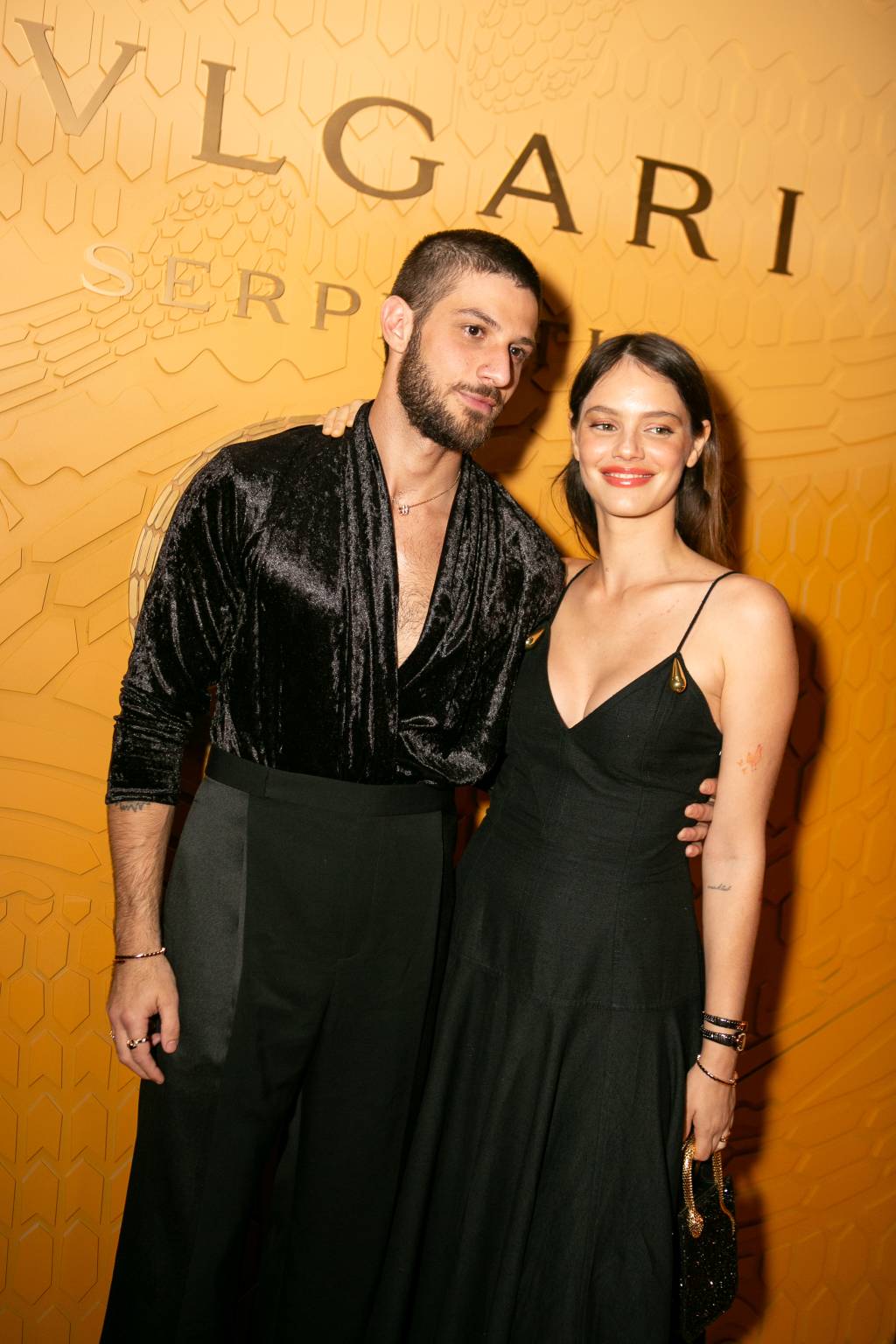 Chay Suede, Laura Neiva Casal posa em frente a parede laranja com a marca Bvlgari. Vestem peças pretas.