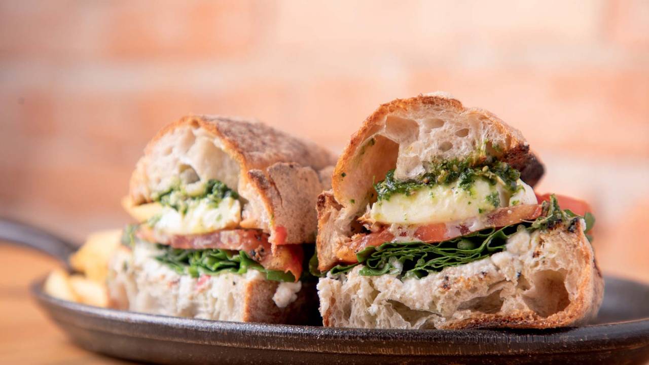Sanduíche fatiado ao meio mostra as camadas do recheio com queijo, tomate e pesto.