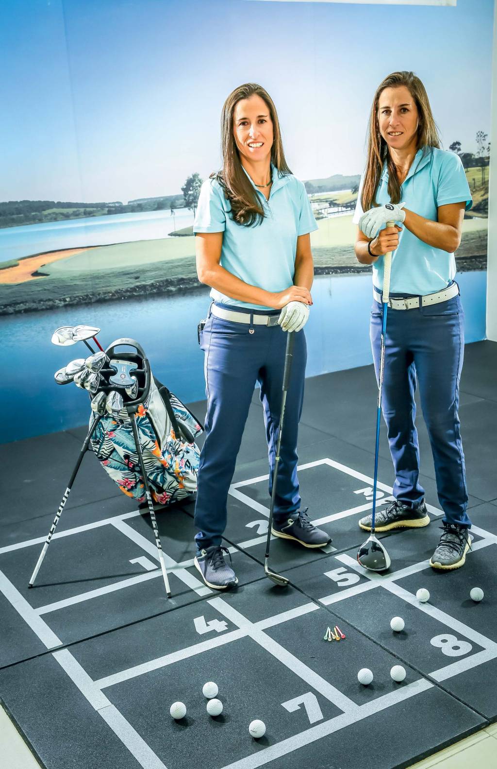 GABRIELA_DANIELAARANTES_GOLF_ (Alexandre Battibugli) Gêmeas de cabelos castanhos lisos posam de pé segurando taco de golfe em ambiente com fundo de tela que imita um campo.
