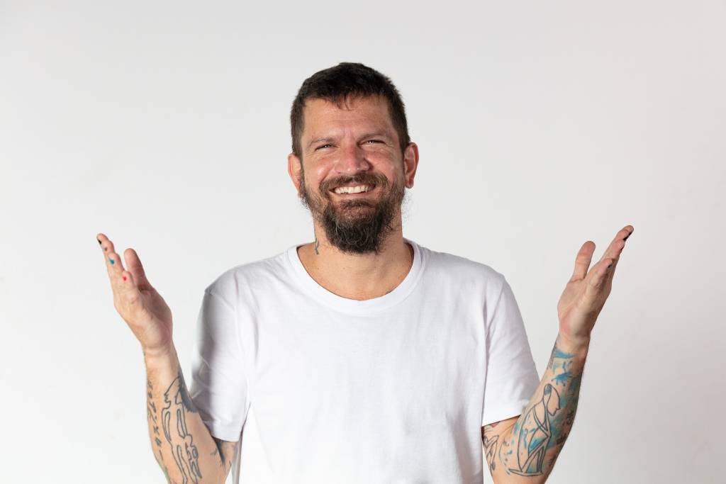 Saulo Fernandes (Sercio Freitas-Divulgação) (2) Homem branco de barba posa com os braços abertos, sorrindo e vestindo camiseta branca.
