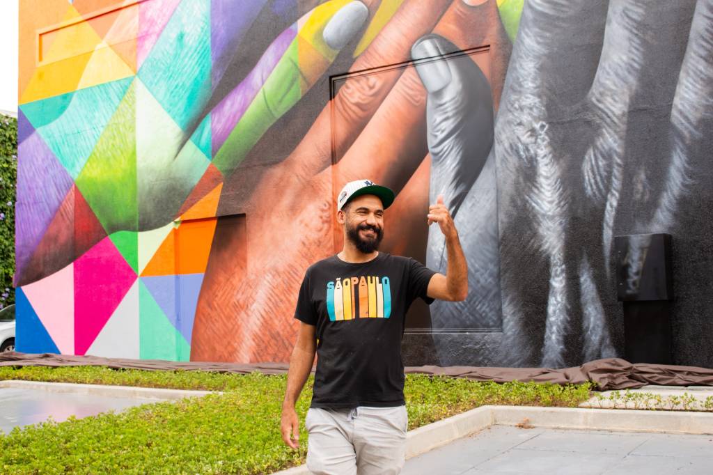 Kobra – Mural EB Park (Carolina Uzueli-Divulgação) (3) Artista com barba, camiseta preta com estampa colorida e shorts brancos posa com uma mão para cima. Ele está em frente a grande mural em área externa, com arte colorida que exibe mãos unidas em vários tons