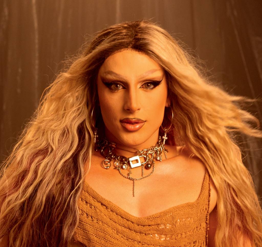 Diego-Martins-Crédito-Catharina-Paiva-Divulgação.jpeg Homem branco em versão drag queen com peruca loura, blusinha marrom e colares dourados