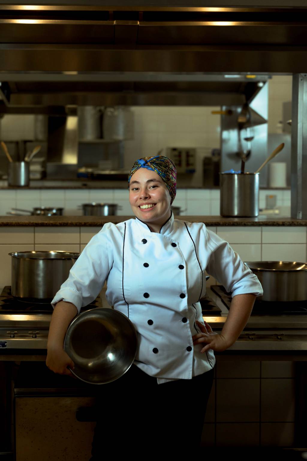 bia azevedo Imagem mostra Bia sorrindo, com roupa de chef de cozinha, segurando um grande recipiente de alumínio na lateral do corpo. Ela está em uma cozinha