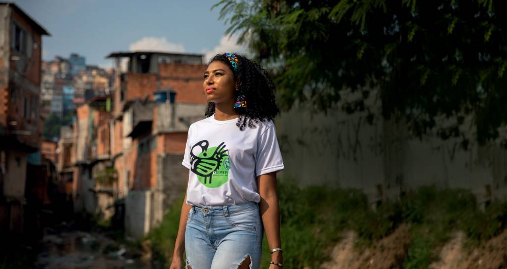 amanda costa Imagem mostra Amanda em pé em rua de bairro da periferia, olhando para o canto esquerdo da foto. Ela usa camiseta branca e calça jeans e uma faixa no cabelo encaracolado