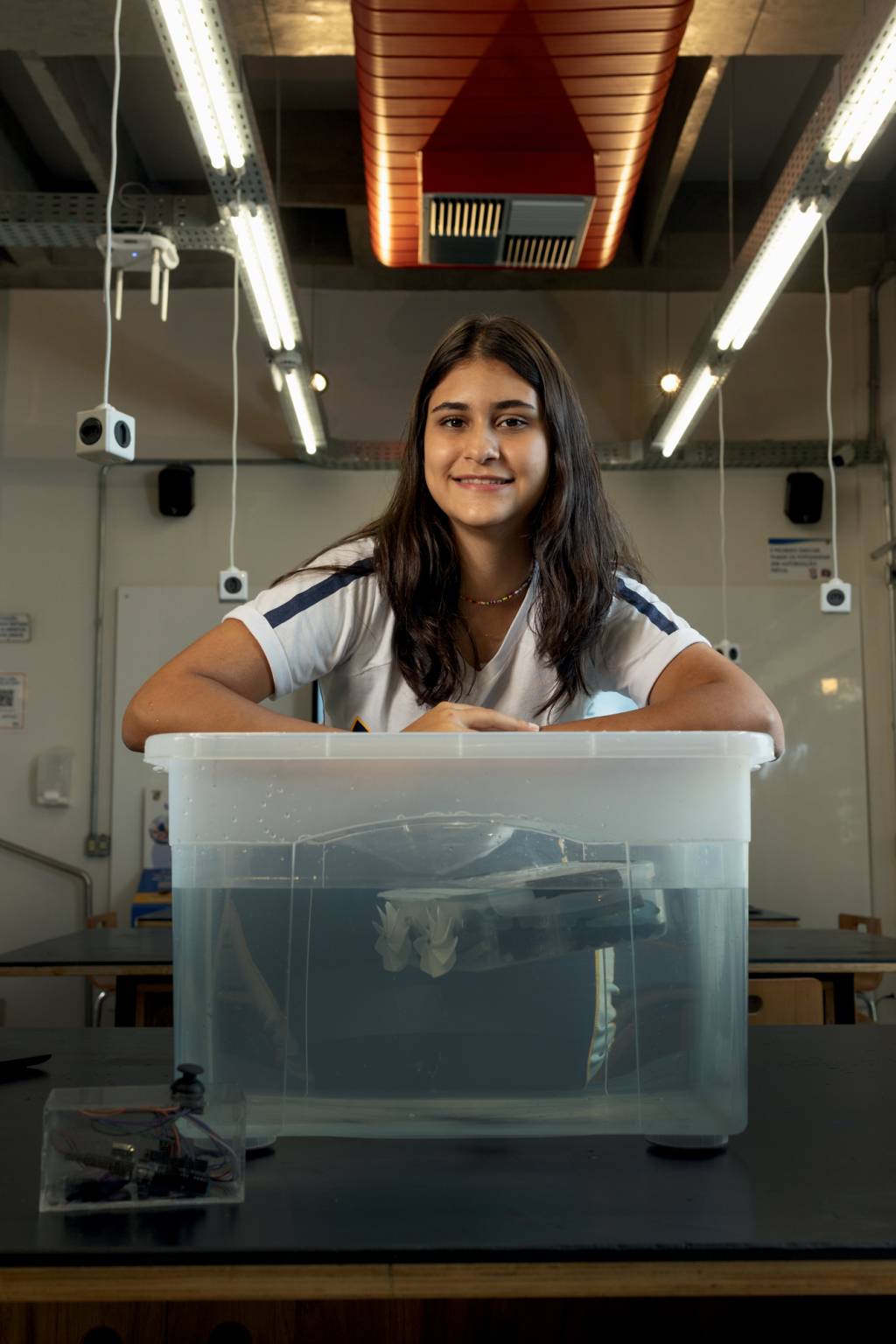 mariana lira Imagem mostra jovem sentada em cima de mesa com uma caixa de plástico transparente cheia de água e com um dispositivo no interior da caixa