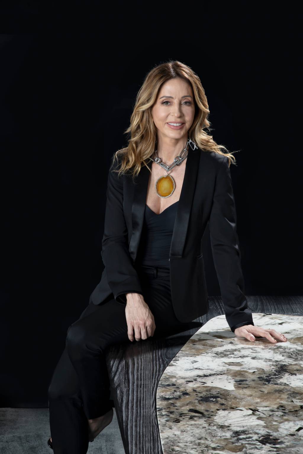 Fernanda Marques-Crédito- Ruy Teixeira-Divulgação Mulher branca com roupas pretas posa sentada em mesa de mármore. Também usa colar grande com pedra amarela e tem cabelos loiros longos.