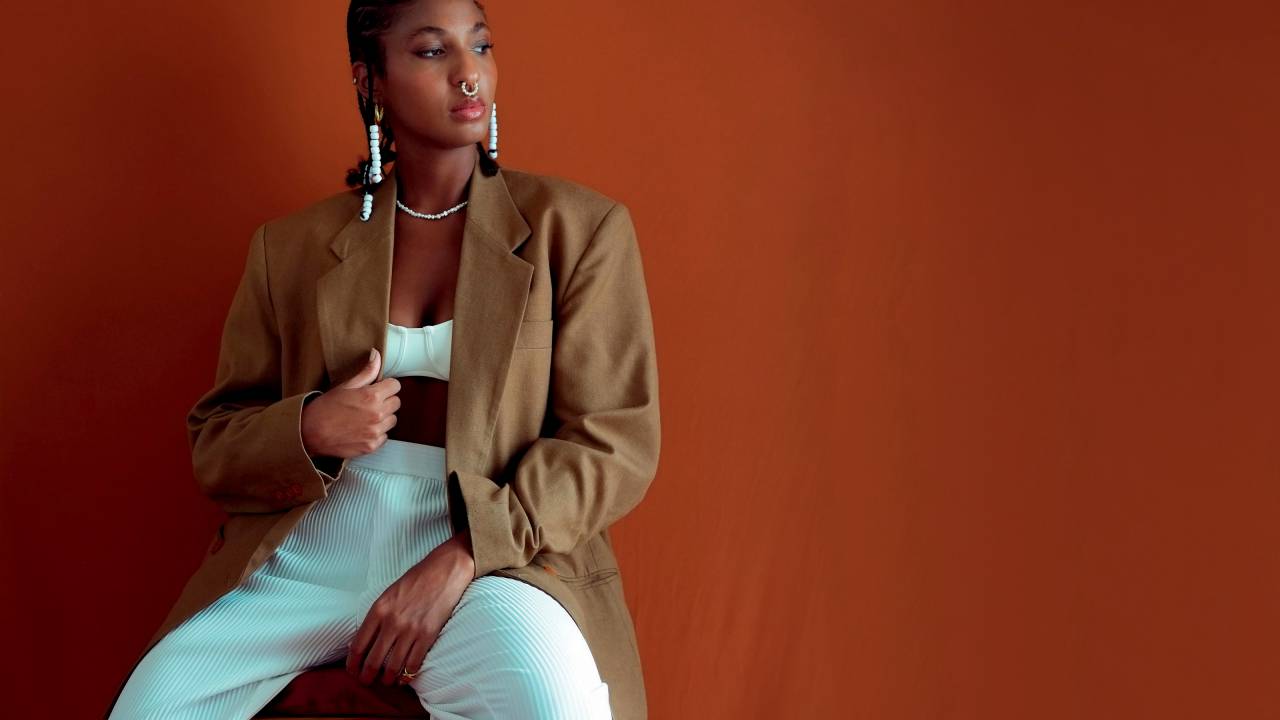 Imagem mostr ajovem sentada em cadeira usando blazer, calça branca e acessórios, olhando para o lado direito em fundo de tom marrom
