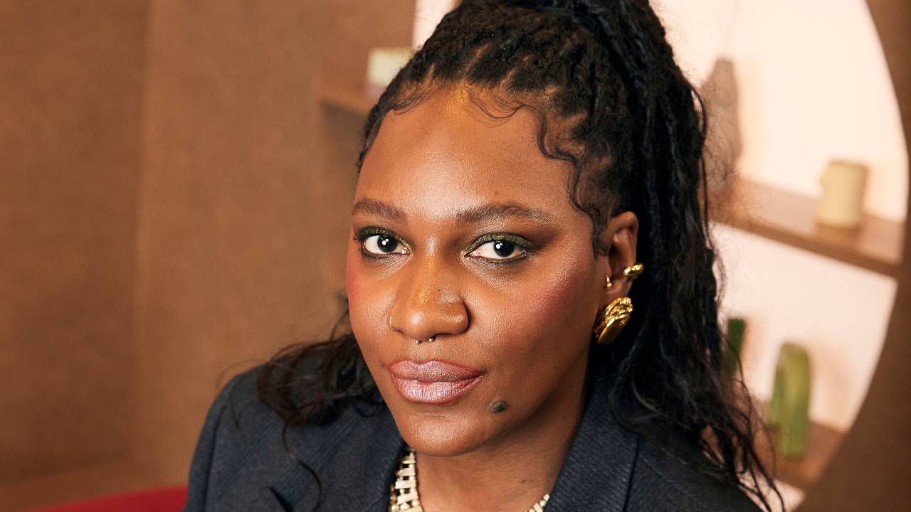 Mulher negra encara a câmera sentada vestindo blazer preto e colar dourado
