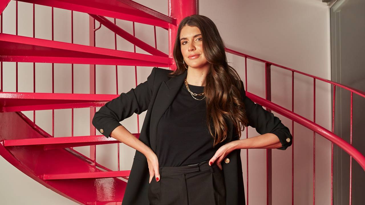Manuela Bordasch posa apoiada em escada vermelha com mãos na cintura. Tem cabelos longos castanhos e veste blazer e camiseta preta.