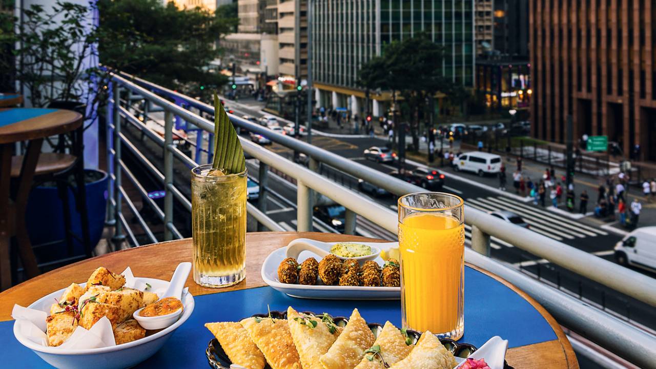 Mesa de madeira em varanda com vista para a Avenida Paulista com porção de pastel, dois drinques, porção de bolinho sobre a mesa.