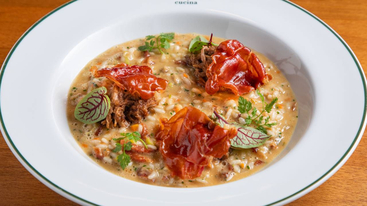Prato fundo de louça branca com risoto coberto por pedaços de presunto cru e folhinhas.