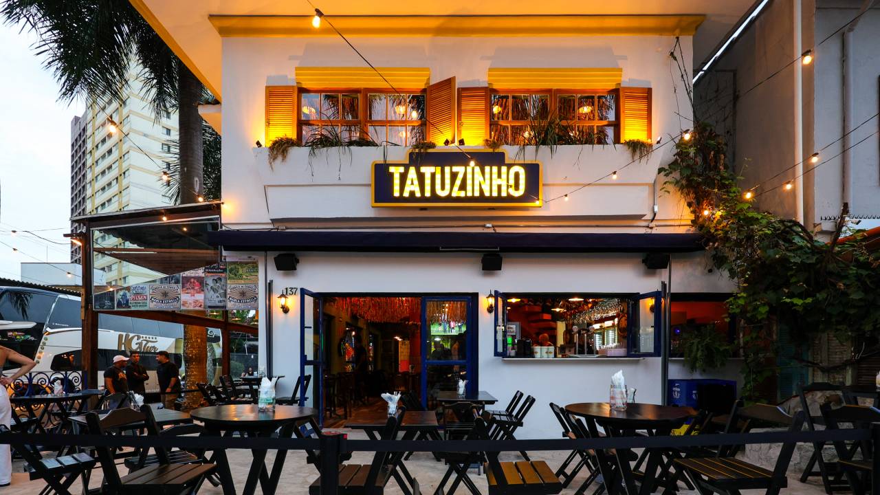 Fachada do bar Tatuzinho, com mesas de madeira na frente, parede branca e cordão de luzes.