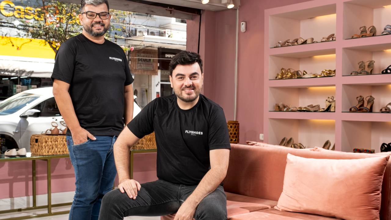 Dois homens posam em loja rosa de sapatos, um sentado e outro em pé
