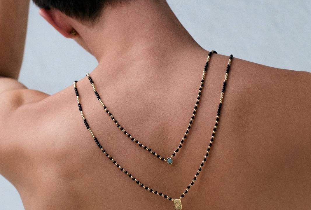 Moço de costas exibe dois colares, um com pedra na ponta e outra com plaquinha de ouro