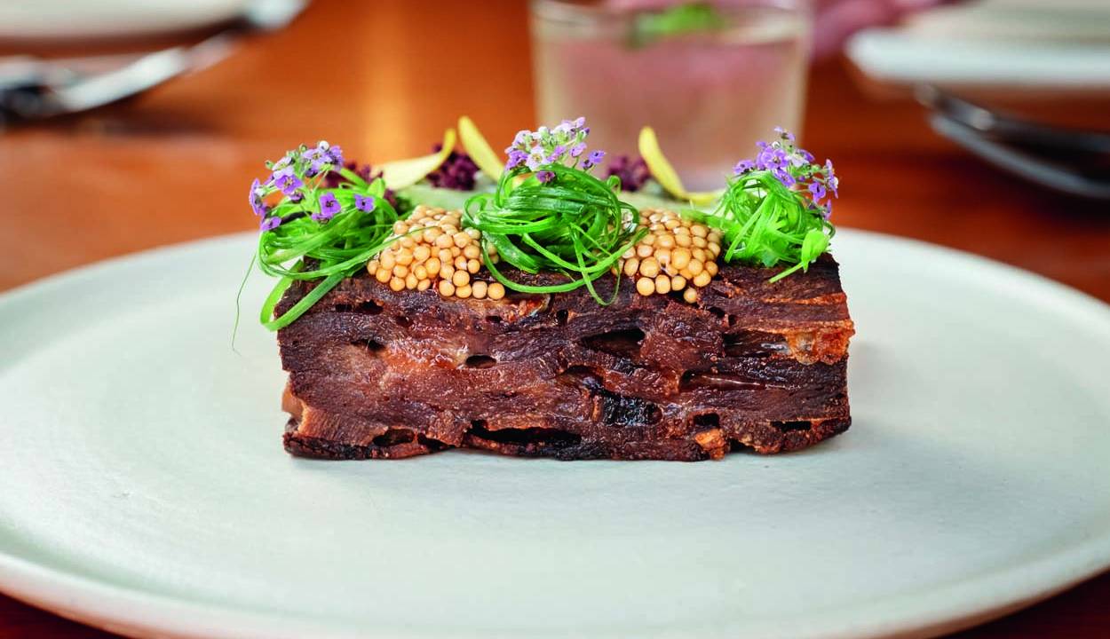Prato de louça branca abaixo de retângulo de carne coberto por grãos de mostarda e fios de folhas verdes.