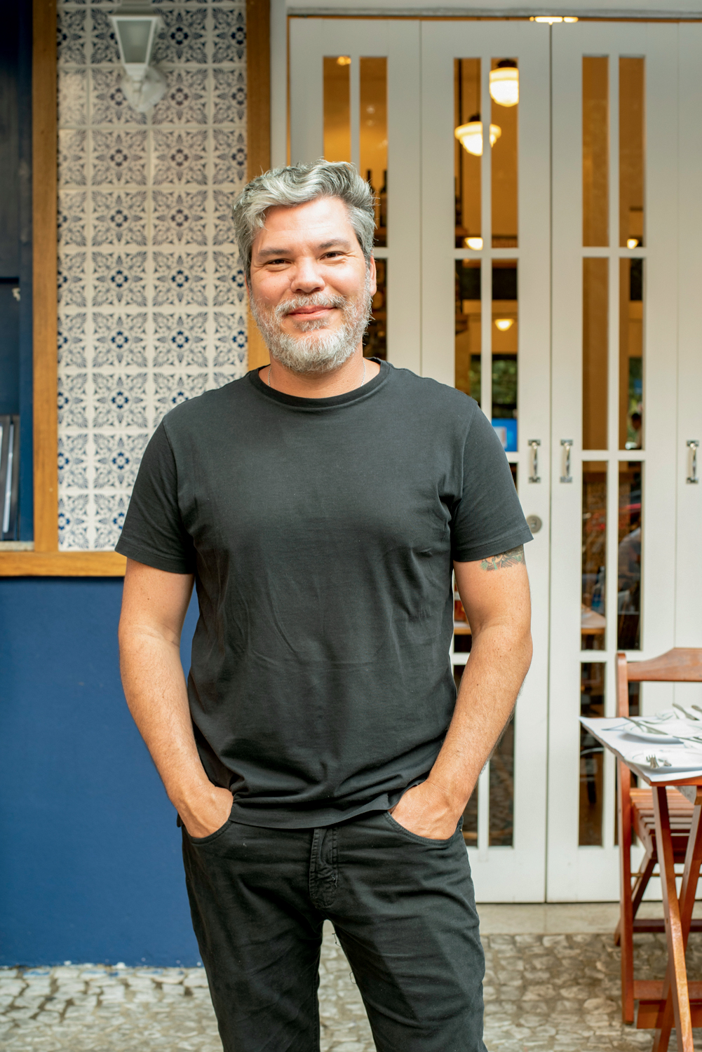 Pedro de Artagão Homem branco de calça e camiseta pretas com as mãos no bolso sorrindo para a foto.