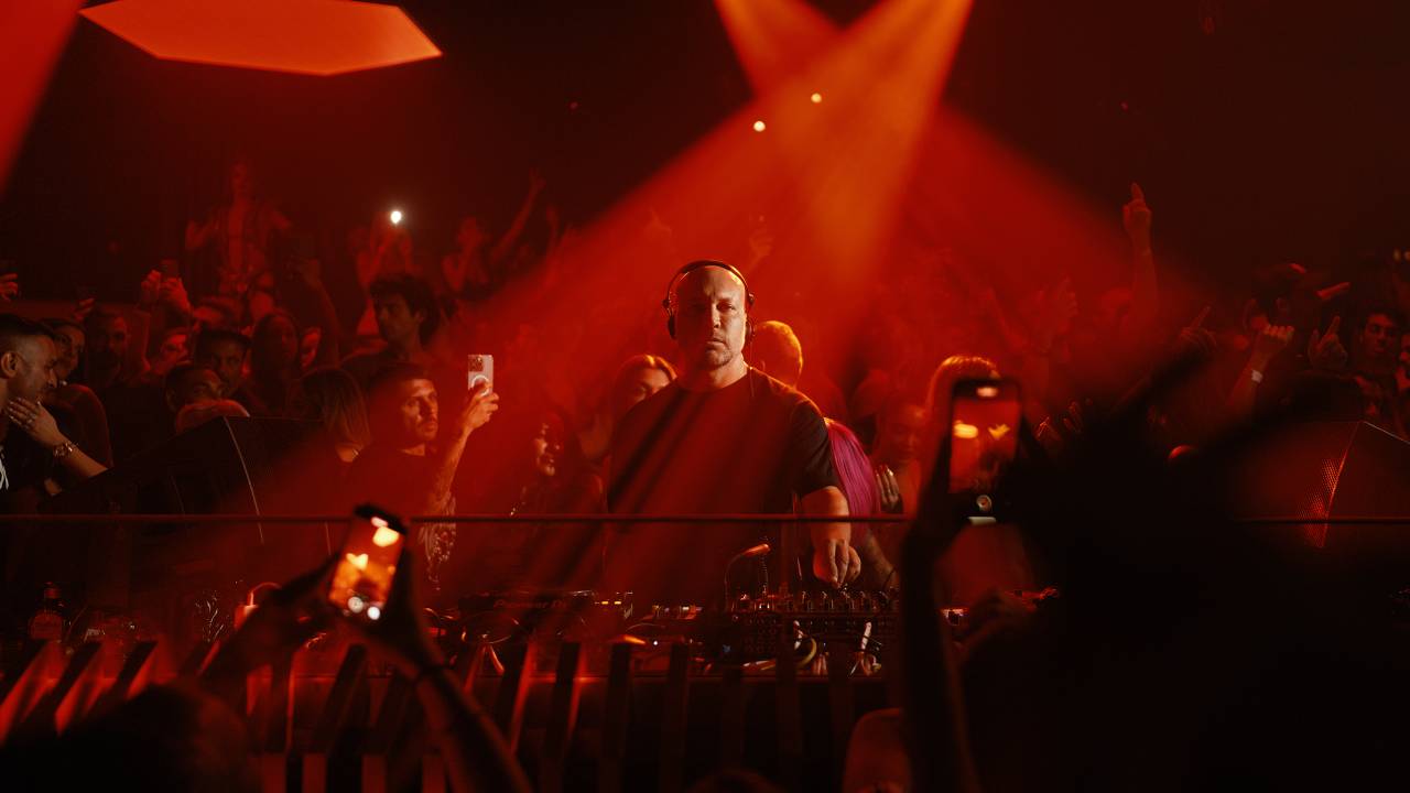 DJ careca com fones em meio a multidão iluminada em tom vermelho