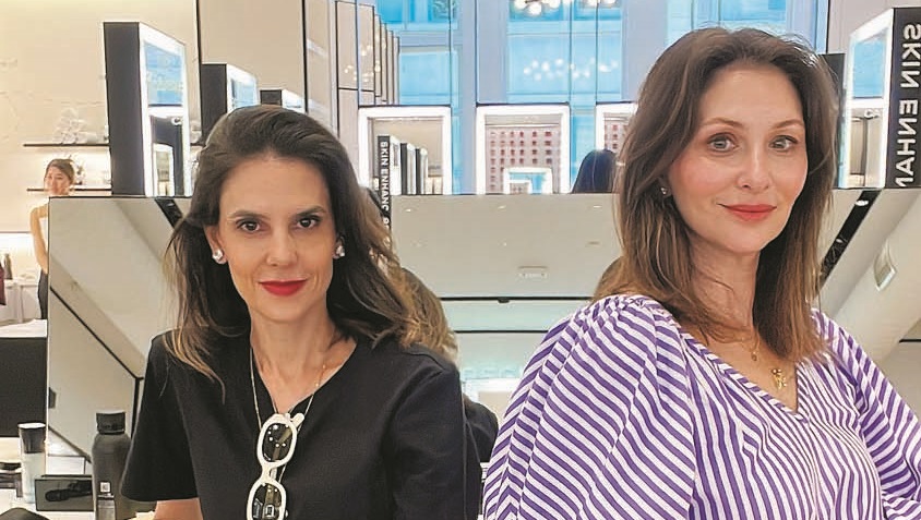 Duas mulheres sentadas lado a lado, a da direita com roupa cor de lavanda, a da esquerda com camiseta preta e calça cinza