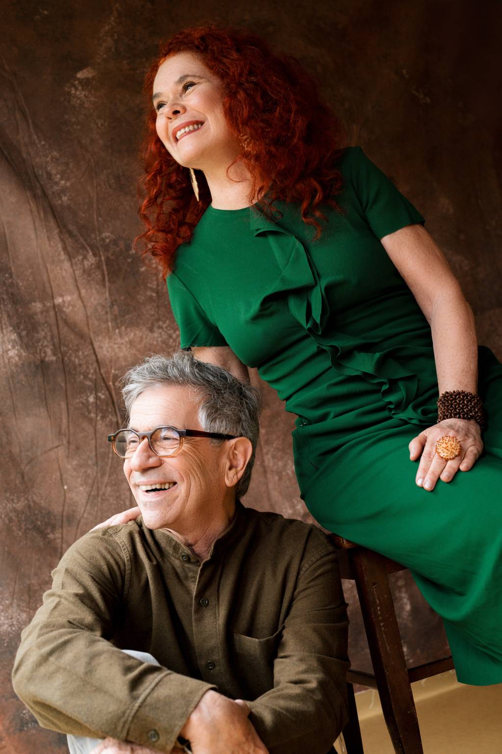 Palavra Cantada Crédito Luana Santos-Estúdio Luana e Mario Fotografia-Divulgação (1) Homem grisalho de óculos sentado e mulher ruiva de vestido verde sentada mais acima, ambos sorrindo e olhando para a esquerda