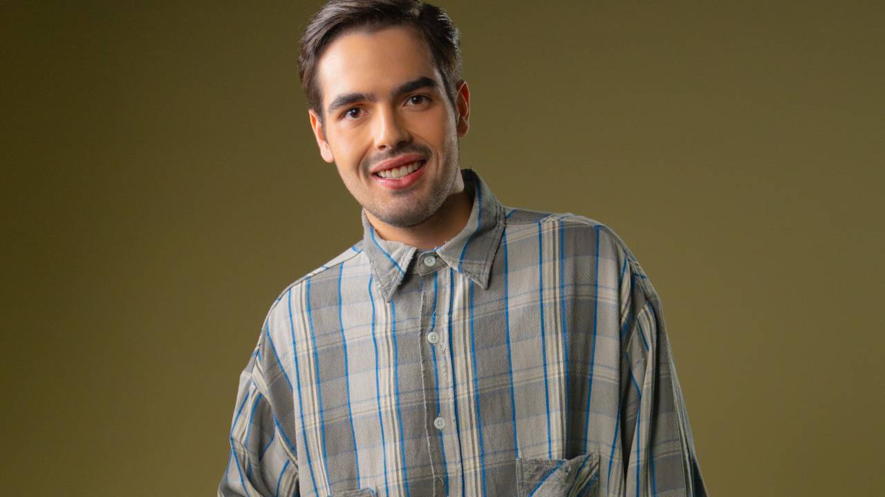 Homem branco de topete e cabelo castanho sorri e veste camisa cinza quadriculada