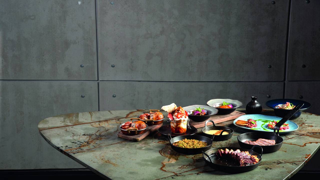 Mesa de mármore cinza esverdeado com menu degustação completo por cima concentrado mais ao lado esquerdo. Com panelinha de ferro com diferentes tipos de carnes e guarnições.